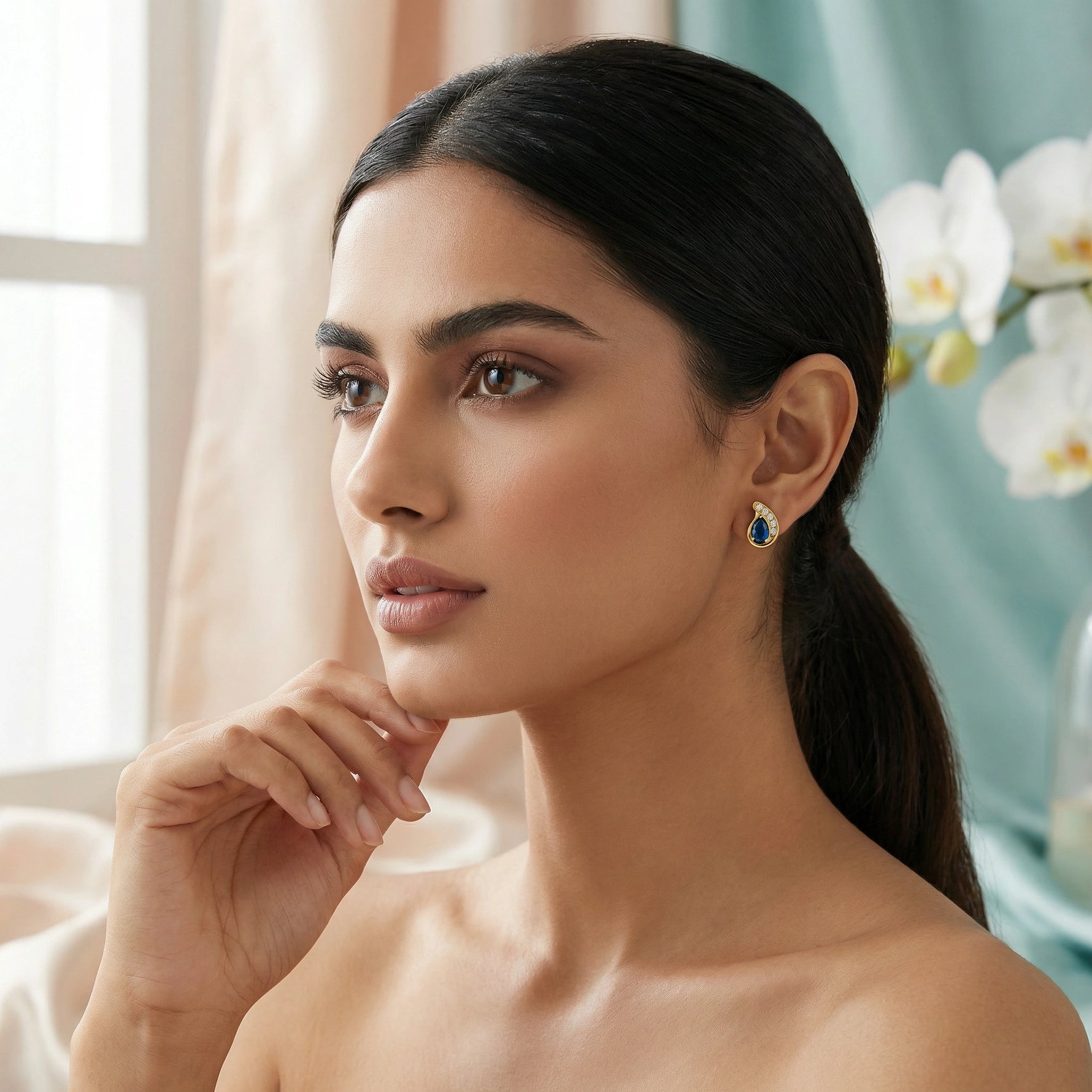 Woman wearing a blue 18k gold plated earring in a softly lit room with flowers in the background
