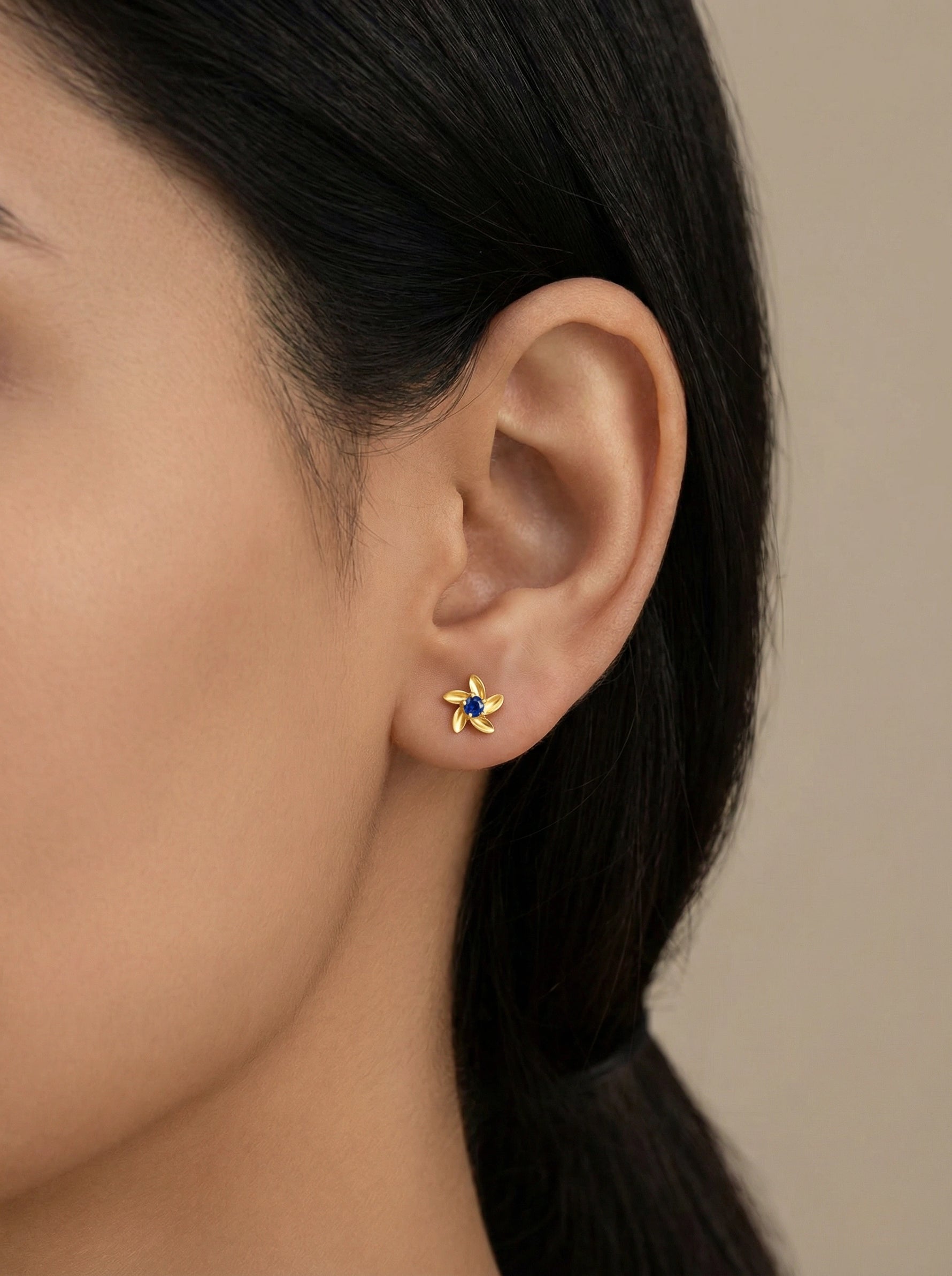Close-up of an ear wearing a 18k gold plated earring with a blue gemstone on a neutral background