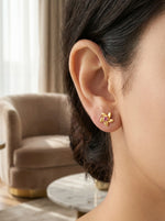 Close-up of a person wearing 18k  gold plated floral earrings with a blurred indoor background.
