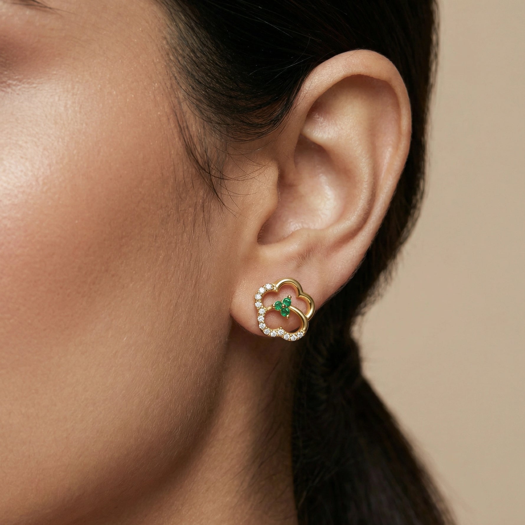 Close-up of an ear wearing a 18k gold plated earring with a green gemstone on a beige background