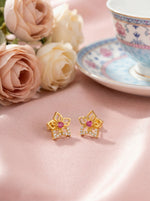 18k Gold plated earrings with a pink gemstone on a pink surface with flowers and a teacup in the background.