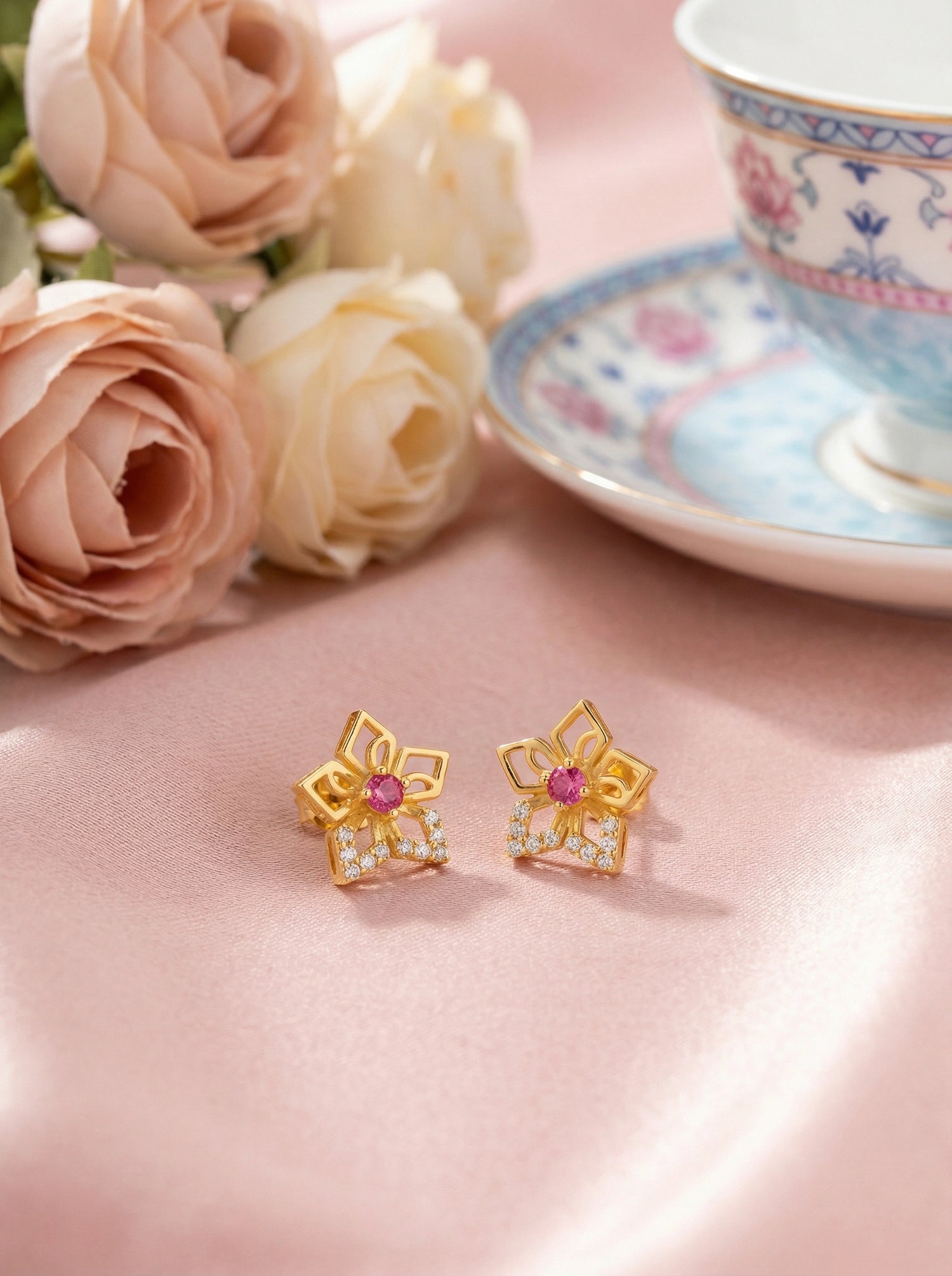 18k Gold plated earrings with a pink gemstone on a pink surface with flowers and a teacup in the background.