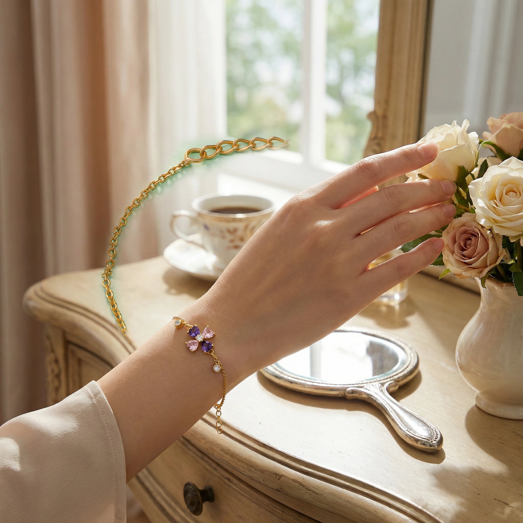 Hand wearing 18k gold plated bracelet with a decorative clasp, reaching towards a cup of coffee and flowers on a table.