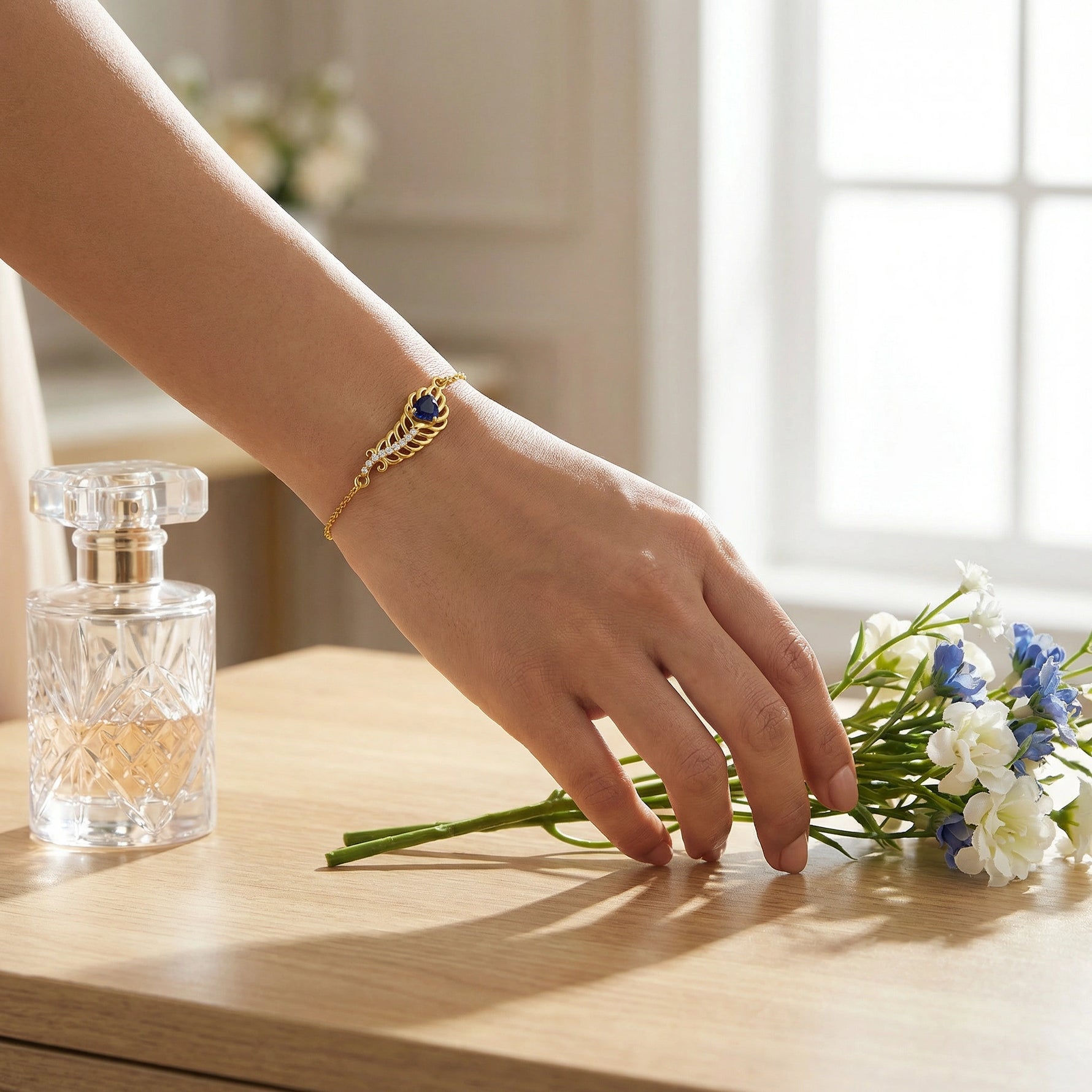 Hand wearing a  18k gold plated bracelet with a blue stone, holding flowers on a wooden surface with a perfume bottle.