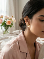 Woman wearing 18k gold plated earrings with a blurred background of flowers and light curtains