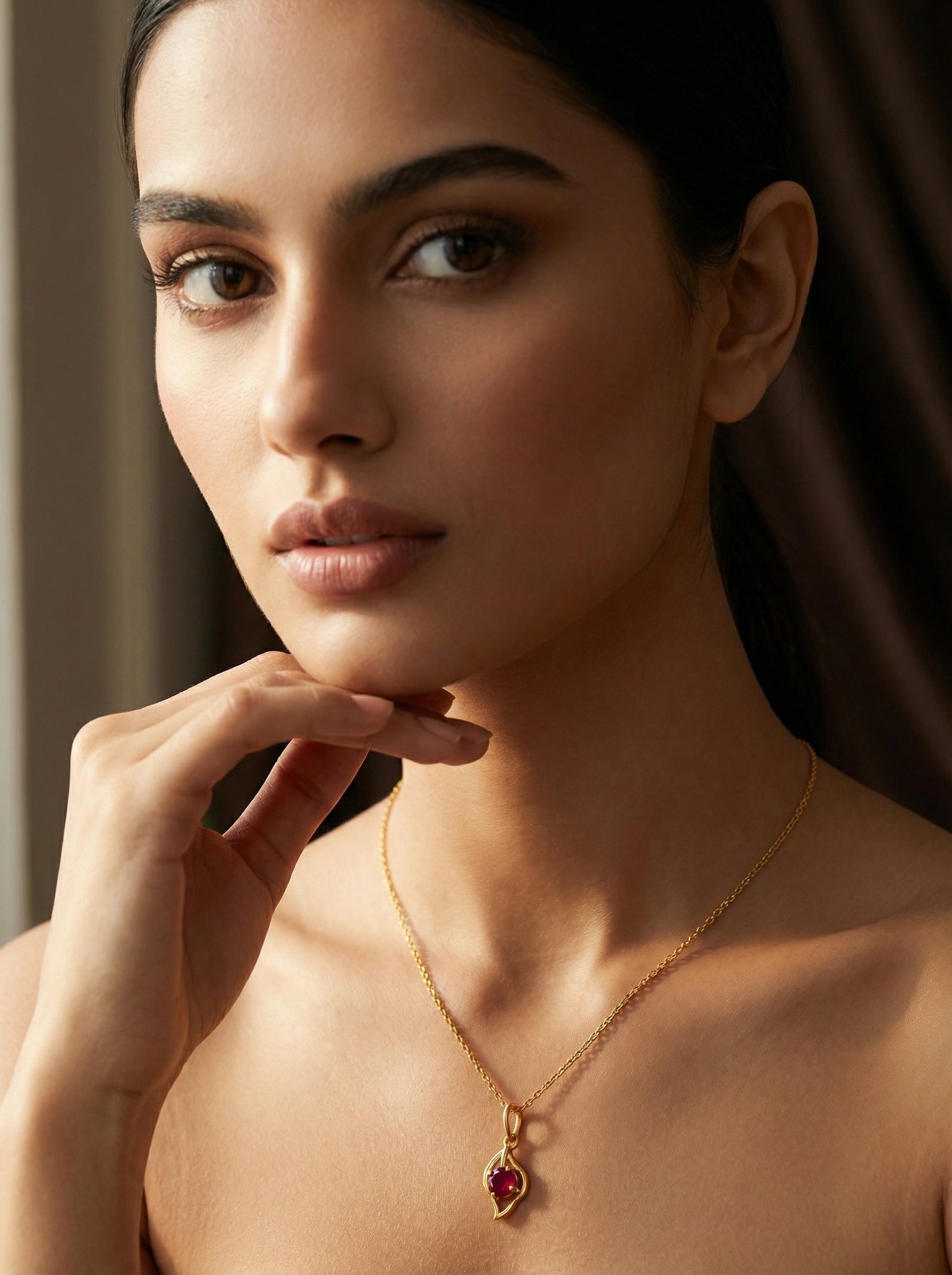 Woman wearing a  18k gold plated necklace with a red gemstone pendant, posing with hand by her face.
