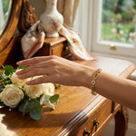 Hand with 18k  gold plated  bracelet reaching towards a bouquet of white roses on a wooden surface.